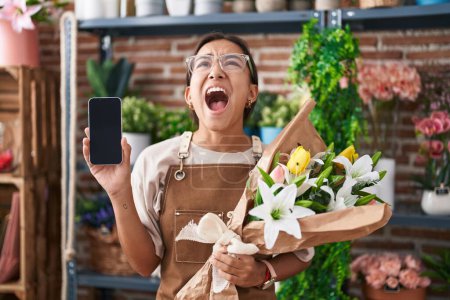 Photo for Young hispanic woman working at florist shop showing smartphone screen angry and mad screaming frustrated and furious, shouting with anger looking up. - Royalty Free Image