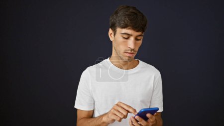 Photo for Cool young hispanic man engrossed in avidly texting a serious message online. he uses smartphone technology with concentrated expression, isolated against black background wall, lifestyle portrait. - Royalty Free Image