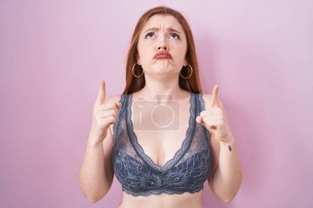 Photo for Redhead woman wearing lingerie over pink background pointing up looking sad and upset, indicating direction with fingers, unhappy and depressed. - Royalty Free Image