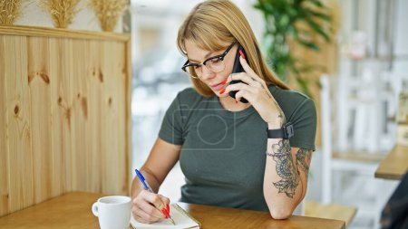 Téléchargez les photos : Jeune femme blonde parlant sur smartphone écrivant sur ordinateur portable au café - en image libre de droit