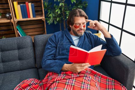 Photo for Middle age man wearing under eye patches reading book on sofa at home - Royalty Free Image