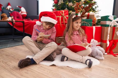 Photo for Brother and sister drawing on notebook sitting on floor by christmas tree at home - Royalty Free Image