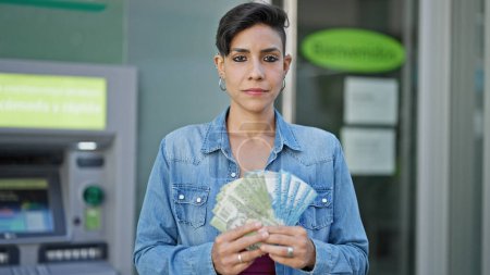 Foto de Joven mujer hispana hermosa sosteniendo billetes chilenos con cara seria en cajero de banco - Imagen libre de derechos