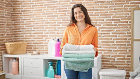 Foto de Joven mujer hispana hermosa sosteniendo toallas dobladas sonriendo en la lavandería - Imagen libre de derechos