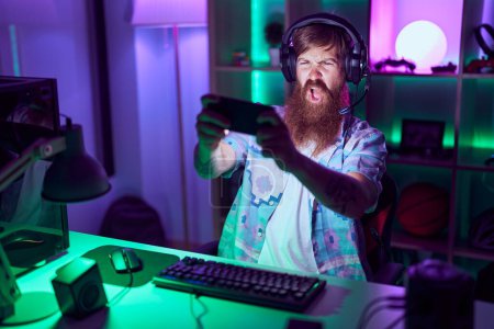 Foto de Young redhead man streamer playing video game using smartphone at gaming room - Imagen libre de derechos