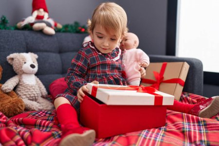 Foto de Adorable chica caucásica desembalaje regalo sentado en el sofá por árbol de Navidad en casa - Imagen libre de derechos