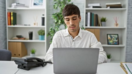 Foto de Atractivo, joven hispano, un profesional exitoso, sentado, enfocado y concentrado en el trabajo en su escritorio de oficina, escribiendo en el portátil con confianza - Imagen libre de derechos
