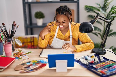 Photo for African american woman with braids sitting at art studio painting looking at tablet smiling happy pointing with hand and finger - Royalty Free Image