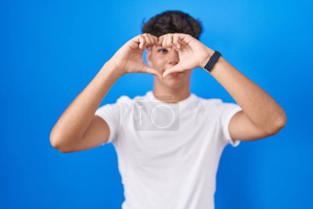 Foto de Adolescente hispana de pie sobre fondo azul haciendo forma de corazón con la mano y los dedos sonriendo mirando a través del signo - Imagen libre de derechos
