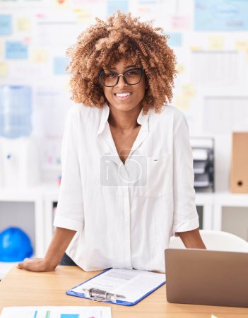 Foto de Trabajadora de negocios afroamericana sonriendo confiada de pie junto al escritorio en la oficina - Imagen libre de derechos