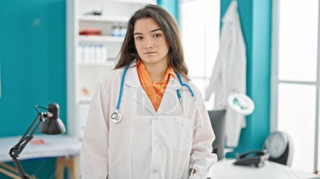 Téléchargez les photos : Jeune belle femme hispanique médecin debout avec une expression sérieuse à la clinique - en image libre de droit