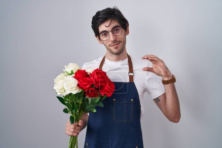 Téléchargez les photos : Jeune homme hispanique tenant bouquet de roses blanches et rouges geste souriant et confiant avec la main faisant signe de petite taille avec les doigts regardant et la caméra. concept de mesure. - en image libre de droit