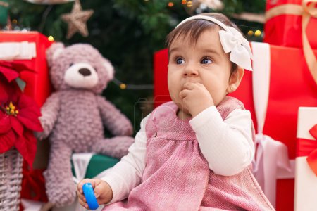 Téléchargez les photos : Adorable jouet hispanique bébé sucer cerceau assis sur le sol par arbre de Noël à la maison - en image libre de droit