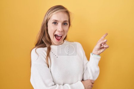 Photo for Young caucasian woman wearing white sweater over yellow background with a big smile on face, pointing with hand finger to the side looking at the camera. - Royalty Free Image