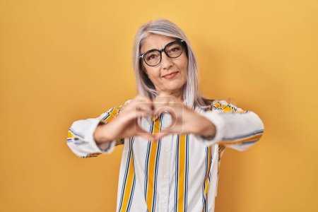 Photo for Middle age woman with grey hair standing over yellow background wearing glasses smiling in love doing heart symbol shape with hands. romantic concept. - Royalty Free Image