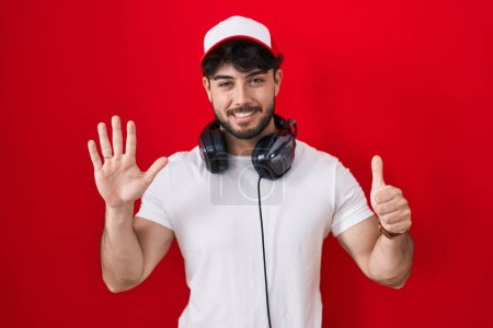 Téléchargez les photos : Homme hispanique avec barbe portant chapeau de joueur et écouteurs montrant et pointant vers le haut avec les doigts numéro six tout en souriant confiant et heureux. - en image libre de droit