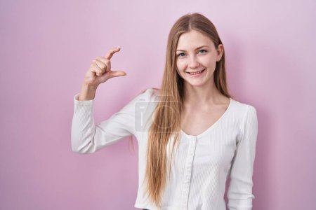Téléchargez les photos : Jeune femme caucasienne debout sur fond rose geste souriant et confiant avec la main faisant signe de petite taille avec les doigts regardant et la caméra. concept de mesure. - en image libre de droit