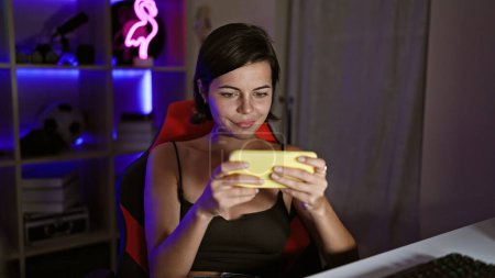 Photo for Confident young hispanic woman, streams smiling and playing video games in home gaming room, using phone - Royalty Free Image