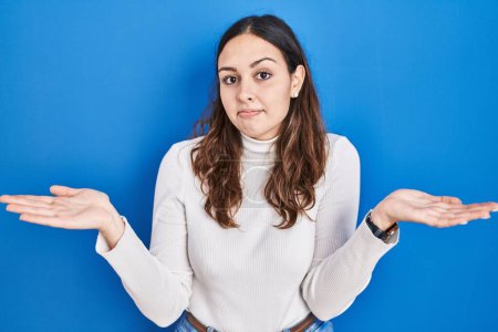 Photo for Young hispanic woman standing over blue background clueless and confused expression with arms and hands raised. doubt concept. - Royalty Free Image