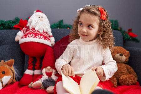 Foto de Adorable chica rubia leyendo libro sentado en el sofá por la decoración de Navidad en casa - Imagen libre de derechos