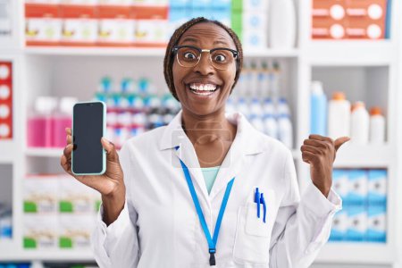 Photo for African american woman working at pharmacy drugstore showing smartphone screen pointing thumb up to the side smiling happy with open mouth - Royalty Free Image