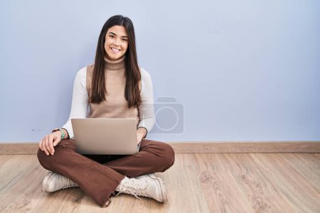 Téléchargez les photos : Jeune femme brune travaillant à l'aide d'un ordinateur portable assis sur le sol avec un sourire heureux et cool sur le visage. chanceux. - en image libre de droit