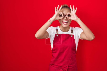 Photo for Young hispanic woman wearing waitress apron over red background doing ok gesture like binoculars sticking tongue out, eyes looking through fingers. crazy expression. - Royalty Free Image