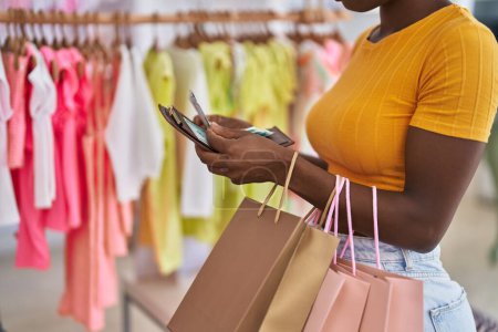 Téléchargez les photos : Femme afro-américaine client tenant des sacs à provisions et portefeuille au magasin de vêtements - en image libre de droit