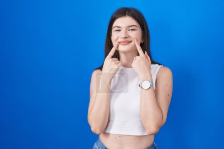 Téléchargez les photos : Jeune femme caucasienne debout sur fond bleu souriant avec bouche ouverte, doigts pointant et forçant sourire joyeux - en image libre de droit