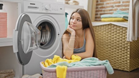 Foto de Joven hermosa chica apoyada en la cesta con la ropa buscando molesto en la sala de lavandería - Imagen libre de derechos
