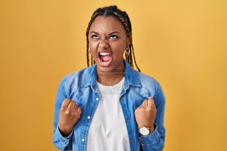 Foto de African american woman with braids standing over yellow background angry and mad screaming frustrated and furious, shouting with anger. rage and aggressive concept. - Imagen libre de derechos