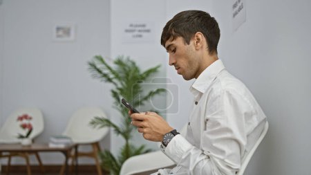 Téléchargez les photos : Sérieux jeune hispanique, un professionnel à succès, assis détendu sur une chaise, absorbé par les textos sur son smartphone, attendant une réunion d'affaires importante dans une salle d'attente spacieuse - en image libre de droit