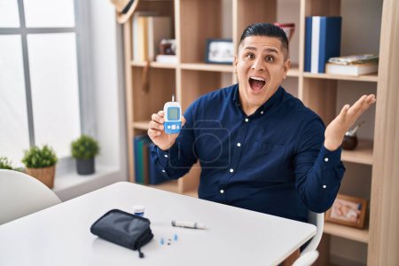 Photo for Hispanic young man holding glucometer device celebrating victory with happy smile and winner expression with raised hands - Royalty Free Image