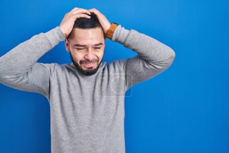 Photo for Hispanic man standing over blue background suffering from headache desperate and stressed because pain and migraine. hands on head. - Royalty Free Image