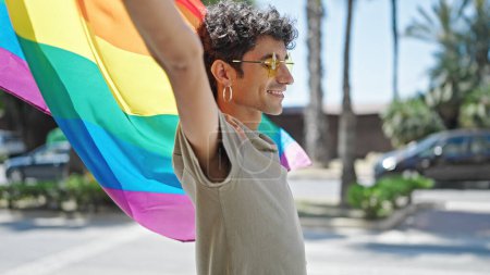Foto de Joven hombre hispano sonriendo confiado llevando bandera de arco iris y gafas de sol en la calle - Imagen libre de derechos