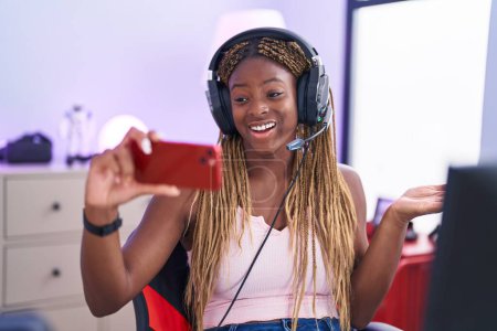 Téléchargez les photos : Femme afro-américaine avec des cheveux tressés jouant à des jeux vidéo avec smartphone célébrant la réalisation avec sourire heureux et expression gagnante avec la main levée - en image libre de droit