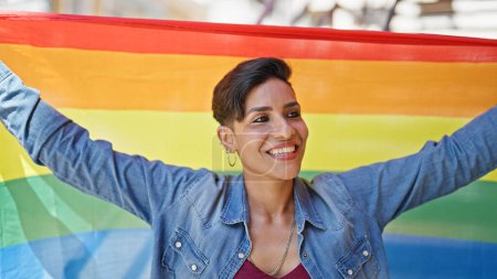 Téléchargez les photos : Jeune belle femme hispanique souriant confiant tenant drapeau arc-en-ciel à la rue - en image libre de droit