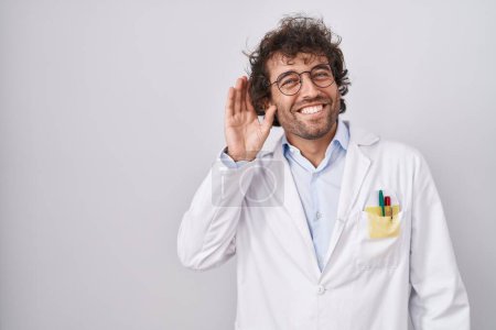 Téléchargez les photos : Jeune homme hispanique portant un uniforme de médecin souriant avec la main sur l'oreille en écoutant une rumeur ou des commérages. concept de surdité. - en image libre de droit