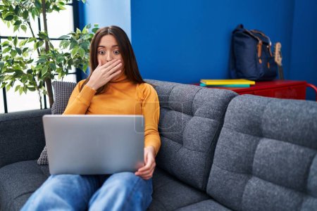 Photo for Young brunette woman working using computer laptop sitting on the sofa covering mouth with hand, shocked and afraid for mistake. surprised expression - Royalty Free Image