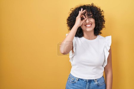 Photo for Young middle east woman standing over yellow background doing ok gesture with hand smiling, eye looking through fingers with happy face. - Royalty Free Image