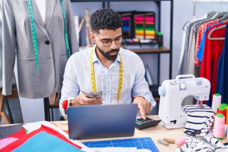 Téléchargez les photos : Jeune tailleur arabe utilisant des dollars de comptage d'ordinateur portable au magasin de tailleur - en image libre de droit