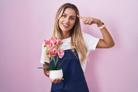 Foto de Mujer rubia joven con delantal de jardinero sosteniendo planta sonriendo apuntando a la cabeza con un dedo, gran idea o pensamiento, buena memoria - Imagen libre de derechos