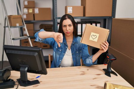 Photo for Hispanic woman working at small business ecommerce holding box with angry face, negative sign showing dislike with thumbs down, rejection concept - Royalty Free Image
