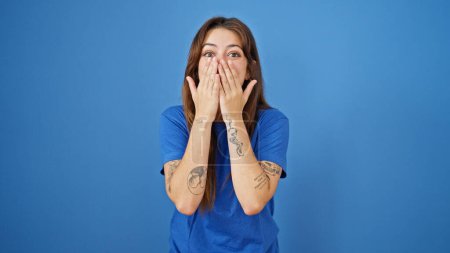 Téléchargez les photos : Jeune belle femme hispanique debout avec expression surprise couvrant la bouche avec les mains sur fond bleu isolé - en image libre de droit