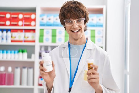Téléchargez les photos : Jeune homme blond pharmacien souriant confiant tenant bouteilles pilules à la pharmacie - en image libre de droit
