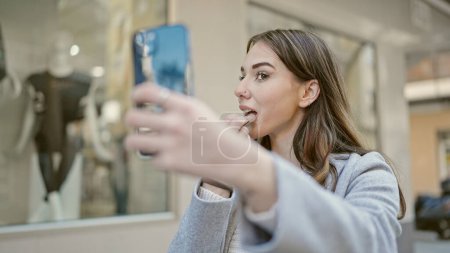 Téléchargez les photos : Jeune femme hispanique utilisant le smartphone comme miroir appliquant du rouge à lèvres dans la rue - en image libre de droit