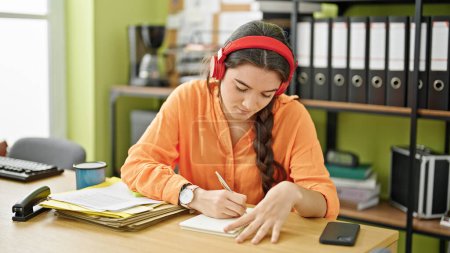 Téléchargez les photos : Jeune belle ouvrière hispanique écoutant de la musique prendre des notes au bureau - en image libre de droit