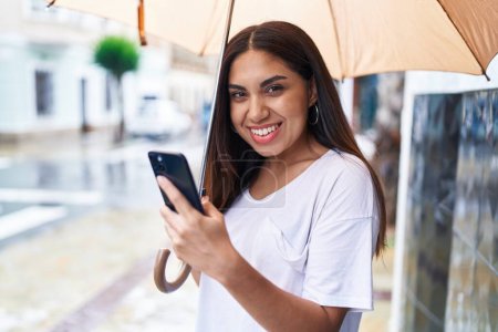 Photo for Young beautiful arab woman using smartphone holding umbrella at street - Royalty Free Image