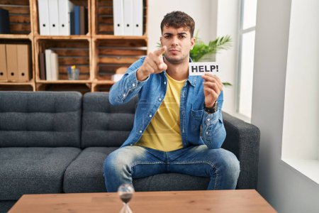 Photo for Young hispanic man at therapy asking for help pointing with finger to the camera and to you, confident gesture looking serious - Royalty Free Image