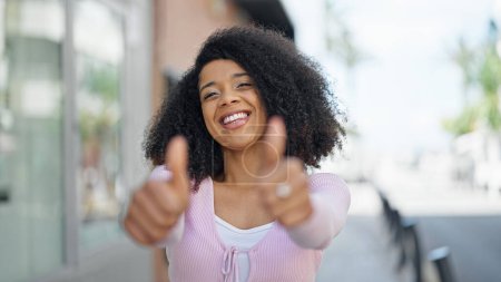 Téléchargez les photos : Afro-américaine femme souriant confiant faire ok signe avec les pouces dans la rue - en image libre de droit